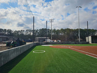 Thumbnail for Tall black backstop netting strung between poles behind the home-plate wall of a turf baseball field, with on-deck circles and a utility cart at left.