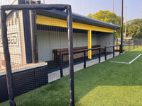 Thumbnail for Black protective netting on padded uprights screening a covered baseball dugout with bench seating, along the turf foul line.