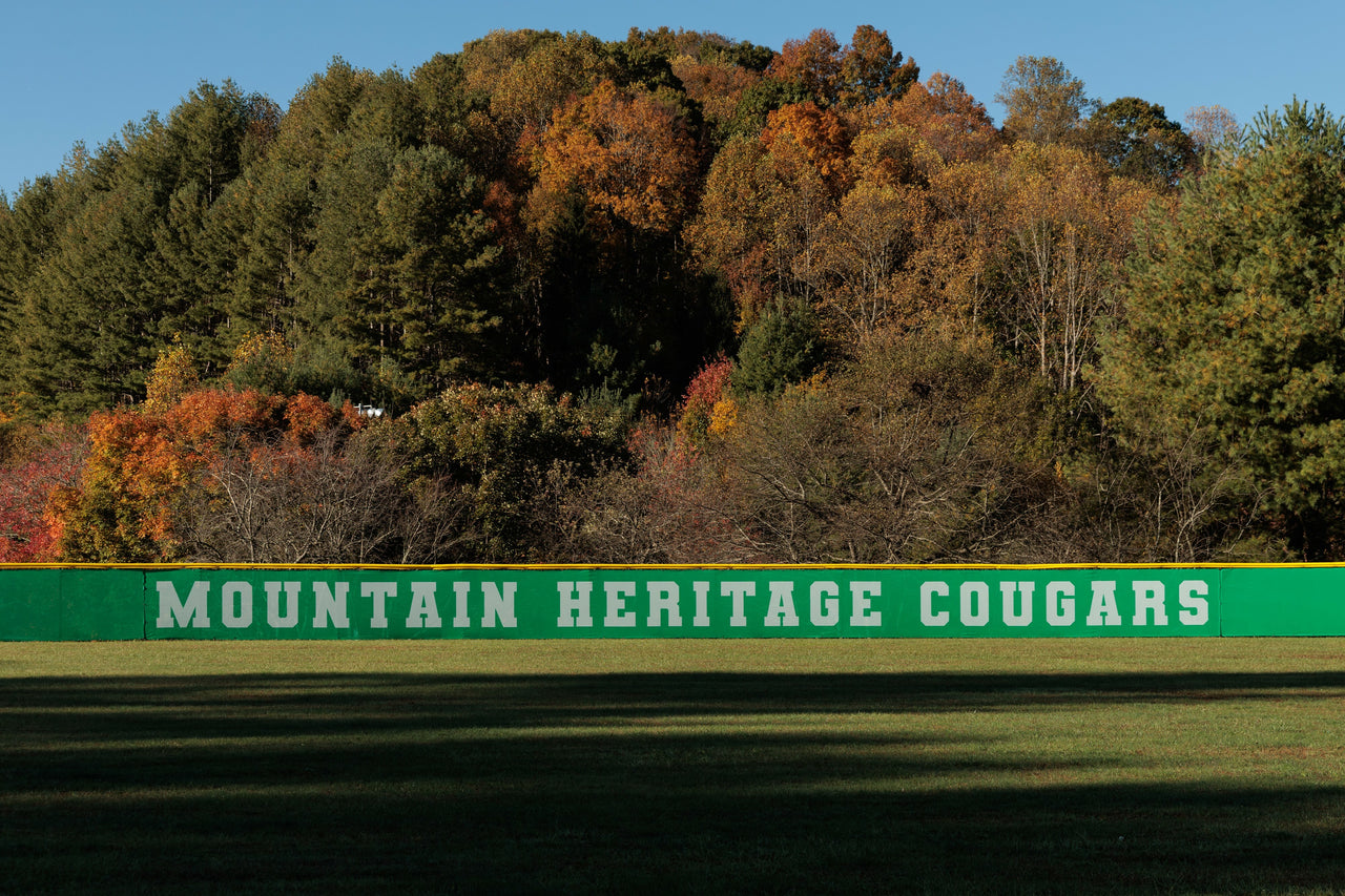 Green printed baseball outfield fence windscreen with Mountain Heritage Cougars team branding along outfield fence