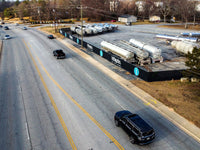 Thumbnail for Aerial view of a custom-branded construction fence screen with the Littlejohn logo enclosing a yard of parked tank trailers beside a road.