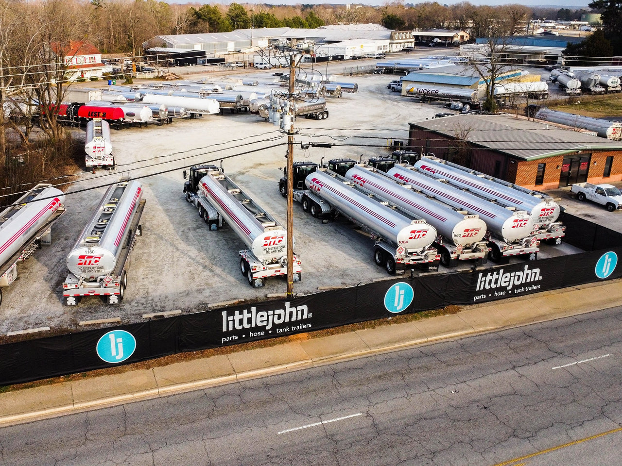 Aerial view of a black construction fence screen printed with the Littlejohn logo, installed along the roadside perimeter fence of a tank-trailer yard.