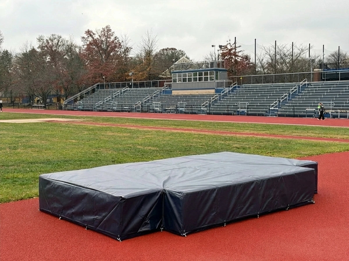 Dark gray weather cover over a high jump mat on a red track in front of stadium bleachers