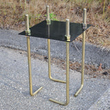 Black square plate with four gold J-shaped anchor bolts and nuts, on asphalt.