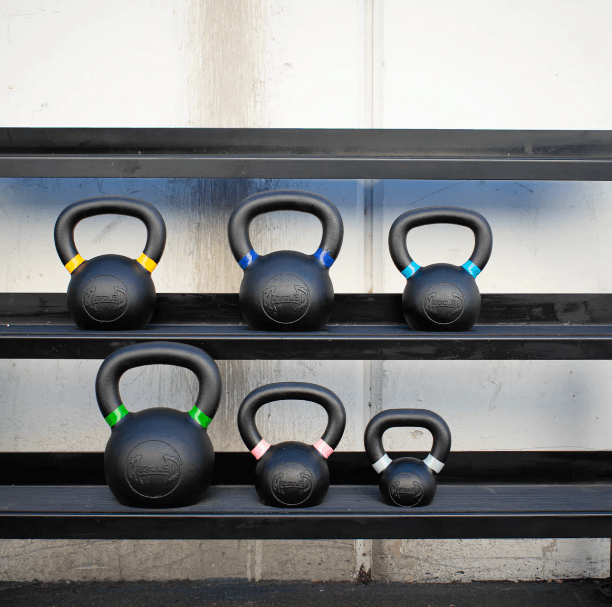 Six black kettlebells with colored handle bands, arranged on two black shelves.