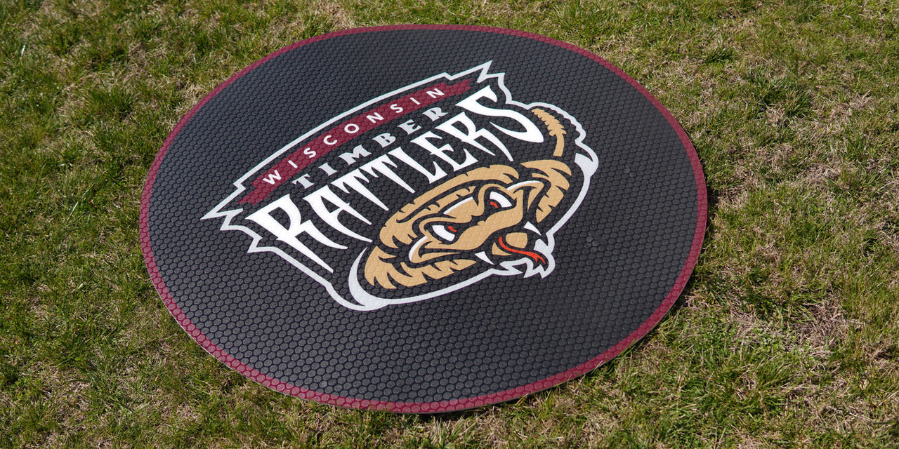 Black circular on-deck circle with "Wisconsin Timber Rattlers" logo on green grass.