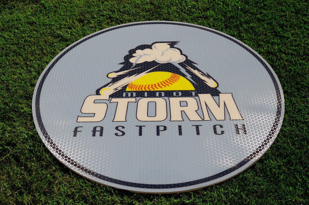 Round gray on-deck circle with "Minot Storm Fastpitch" logo on green grass.