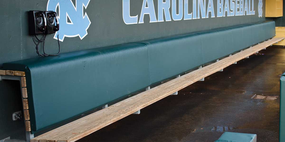 Green padded dugout bench back running the length of a wooden bench below a Carolina Baseball wall