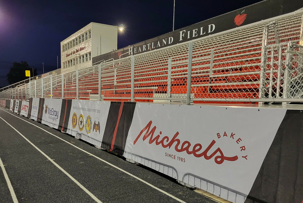Football windscreens printed with Michael's Bakery and other sponsor logos along a stadium track at Heartland Field