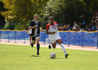 Thumbnail for Blue above-ground fencing with a gold top rail lining a soccer field as an American University player dribbles