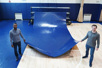 Thumbnail for Two men unrolling a blue vinyl gym floor cover from a storage rack onto a hardwood basketball court
