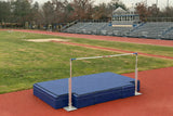 High jump landing mats with standards and bar set up on an outdoor track field beside bleachers and a running track.