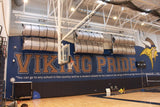 Two rows of gray rolled-up mats on a wall above "Viking Pride" in a school gym.