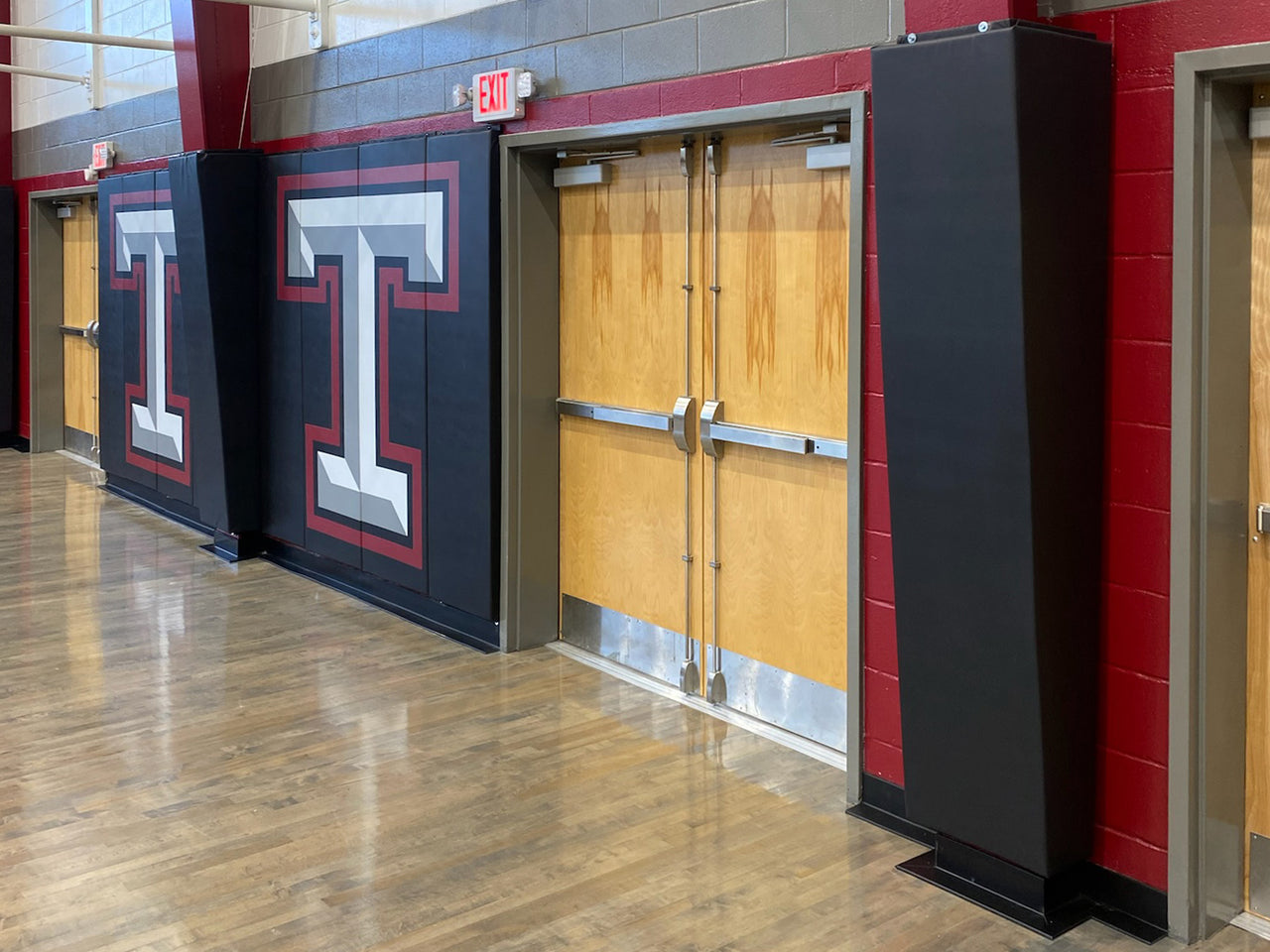 Black gym wall padding printed with large red-outlined white T logos, beside black corner pads and wood doors