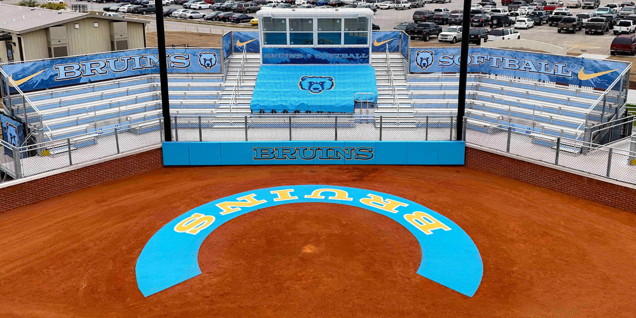 Aerial view of a softball field with blue and white bleachers, a scoreboard, and "BRUINS" written on the field.