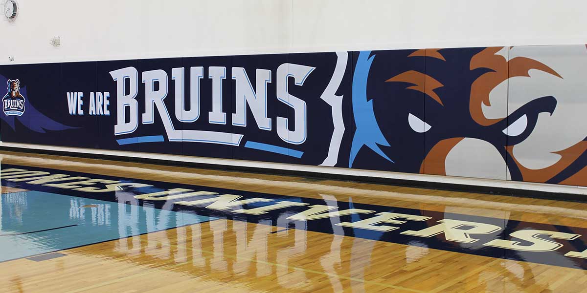 Digitally printed wall panel with "WE ARE BRUINS" text and a bear mascot design in a gym.