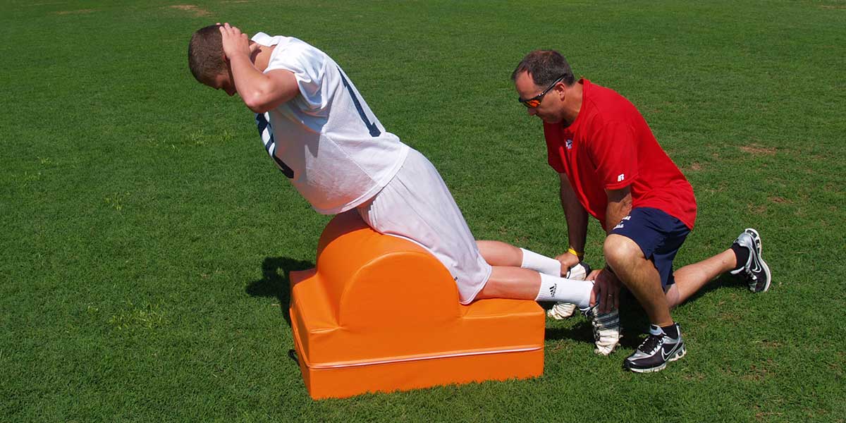 Athlete using an orange glute ham machine on grass, assisted by a coach.