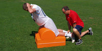 Thumbnail for Athlete using an orange glute ham machine on grass, assisted by a coach.