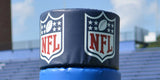 Blue goal post pad cap with two NFL logos, stadium bleachers in background.