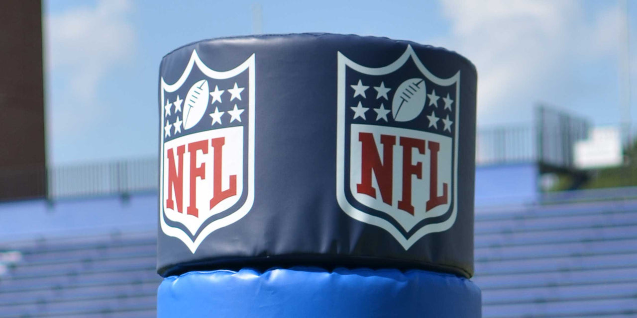 Blue goal post pad cap with two NFL logos, stadium bleachers in background.