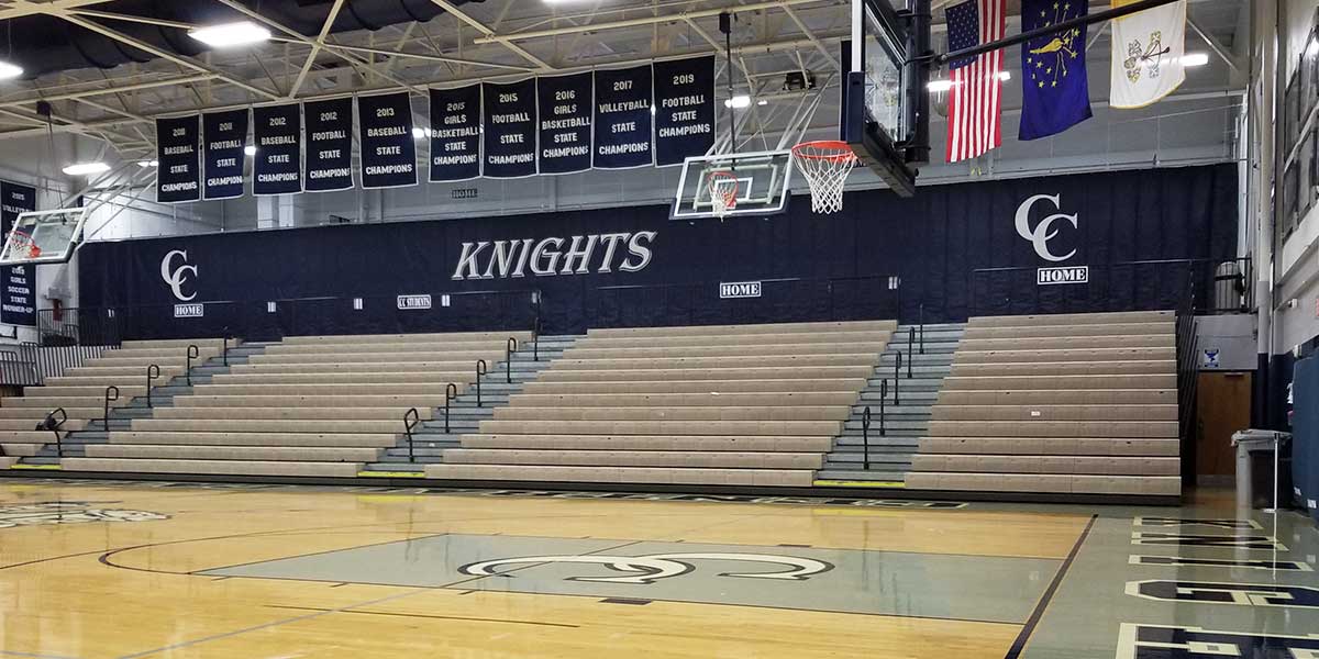 Black gym divider curtain with "KNIGHTS" and "CC HOME" logos above empty bleachers in a basketball court.