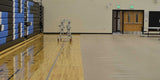 Gym floor partially covered with beige protective mats, revealing hardwood floor and bleachers.