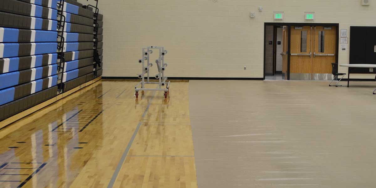 Gym floor partially covered with beige protective mats, revealing hardwood floor and bleachers.