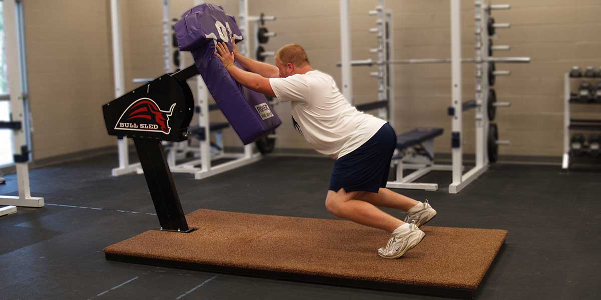 Man pushing a purple football training dummy on a "BULL SLED" base, indoors on a brown mat.