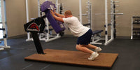 Thumbnail for Man pushing a purple football training dummy on a "BULL SLED" base, indoors on a brown mat.