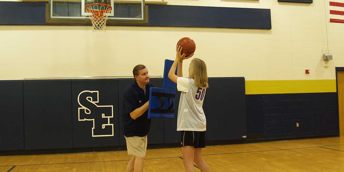 Man holding a blue basketball L shield while a girl shoots a basketball.