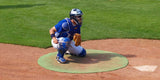 Baseball catcher in blue uniform crouching on a green on-deck circle.