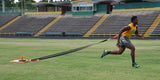 Athlete in a yellow shirt and gray shorts pulls an orange sled across a grassy field.