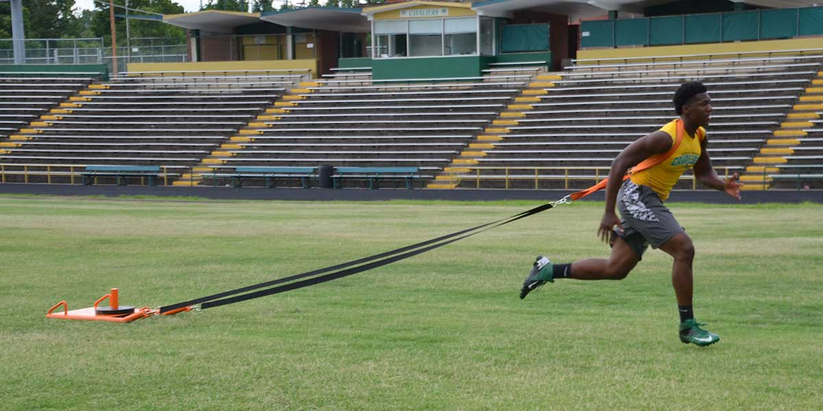 Athlete in a yellow shirt and gray shorts pulls an orange sled across a grassy field.