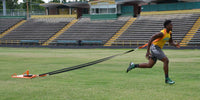 Thumbnail for Athlete in a yellow shirt and gray shorts pulls an orange sled across a grassy field.