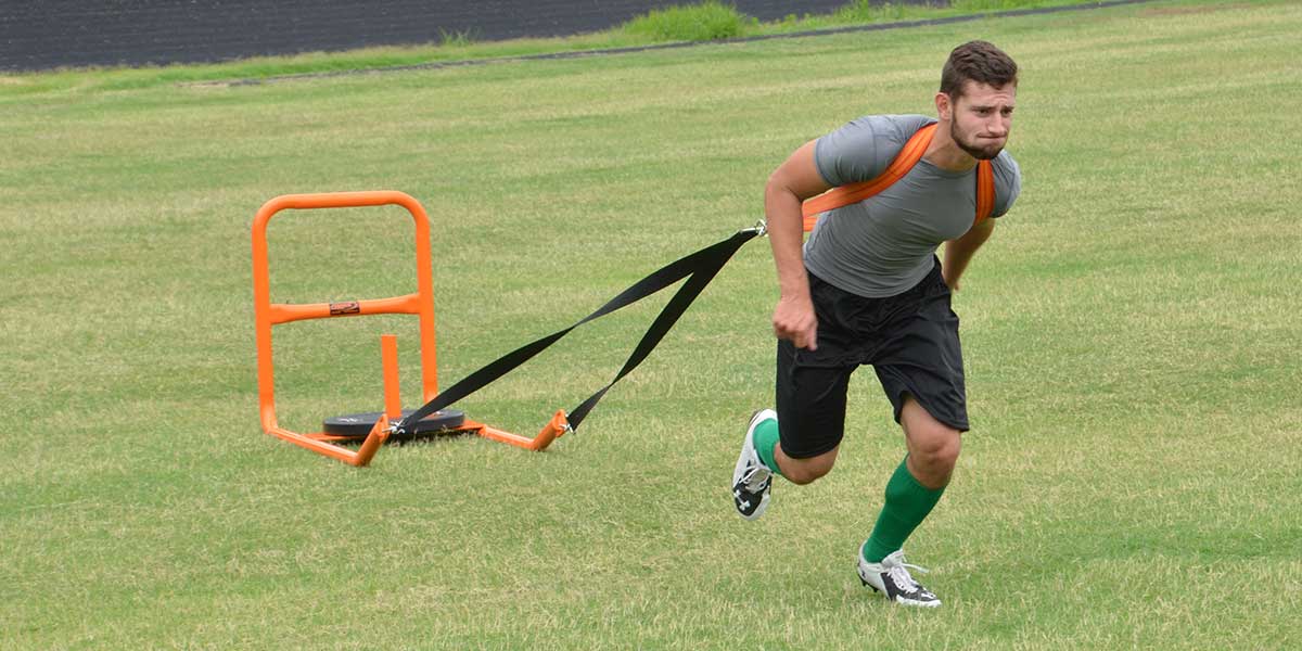 Man in gray shirt and black shorts pulling an orange push-pull sled across a grassy field.