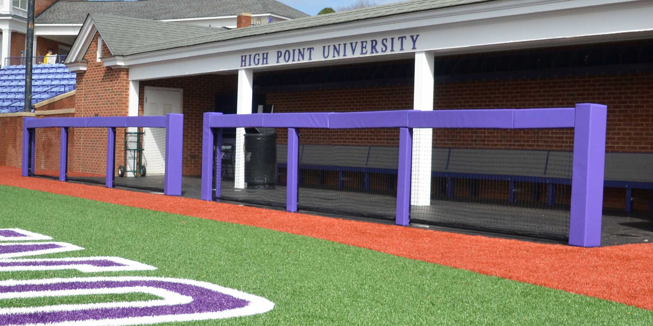 Purple square rail padding on a sports field in front of a brick dugout.