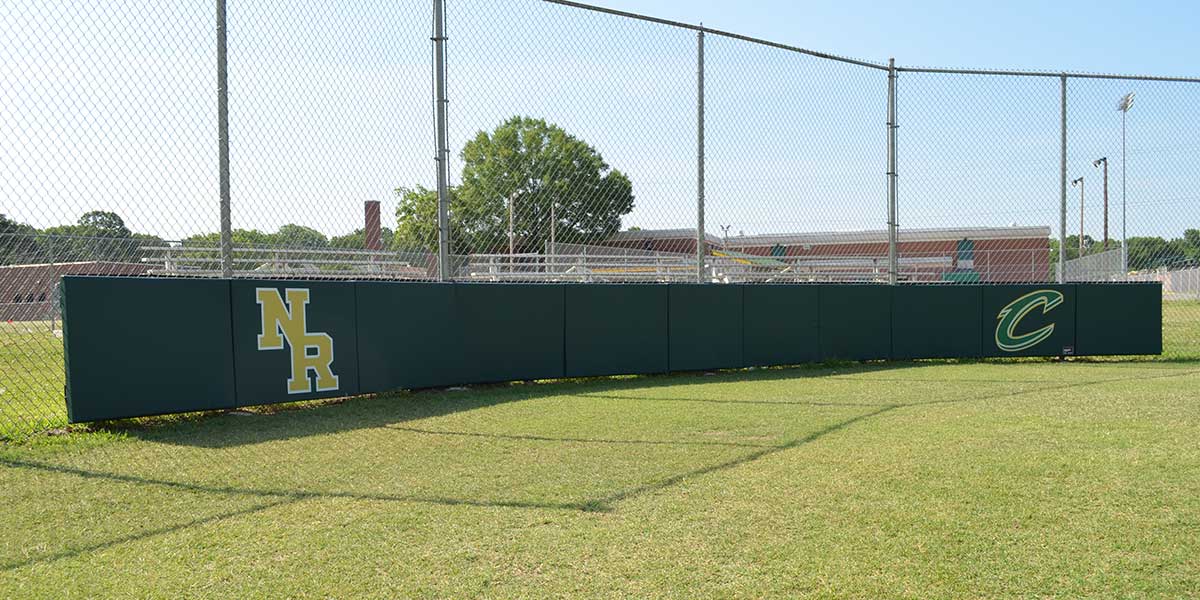 Green wall panels with "NR" and "C" logos installed along a chain-link fence on a baseball field.