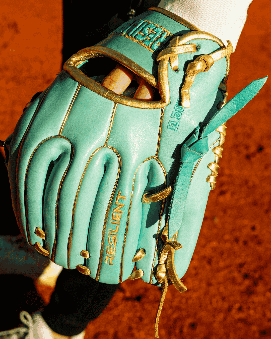 Close-up of a mint green baseball glove with gold trim and "RESILIENT" embroidered on the palm.