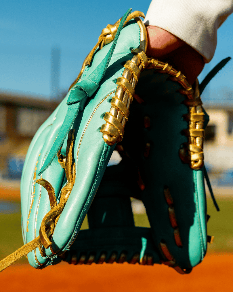 Mint green baseball glove with gold lacing, held by a person, on a sunny baseball field.