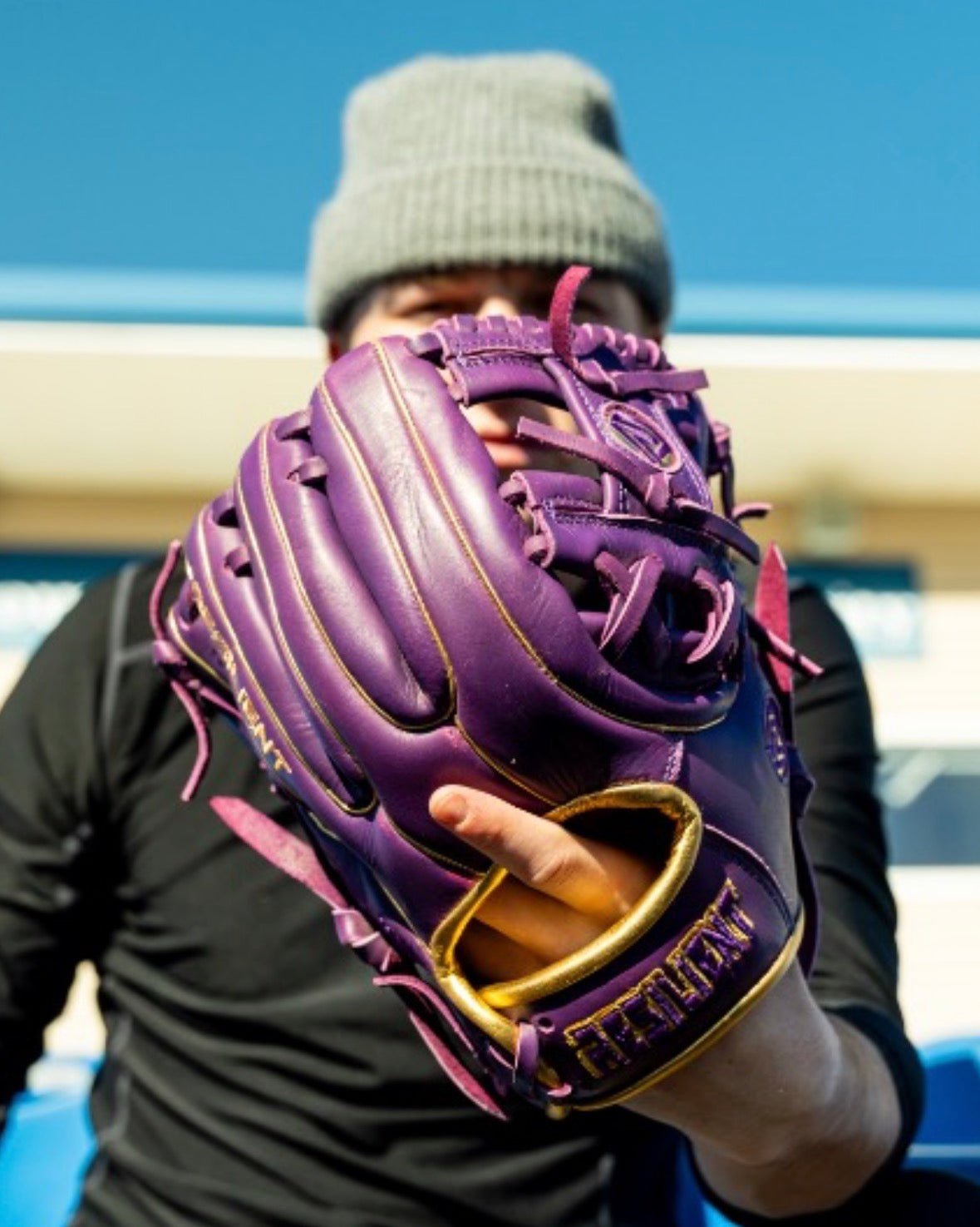 Person wearing a purple and gold baseball glove, obscuring their face, with a blue sky in the background.