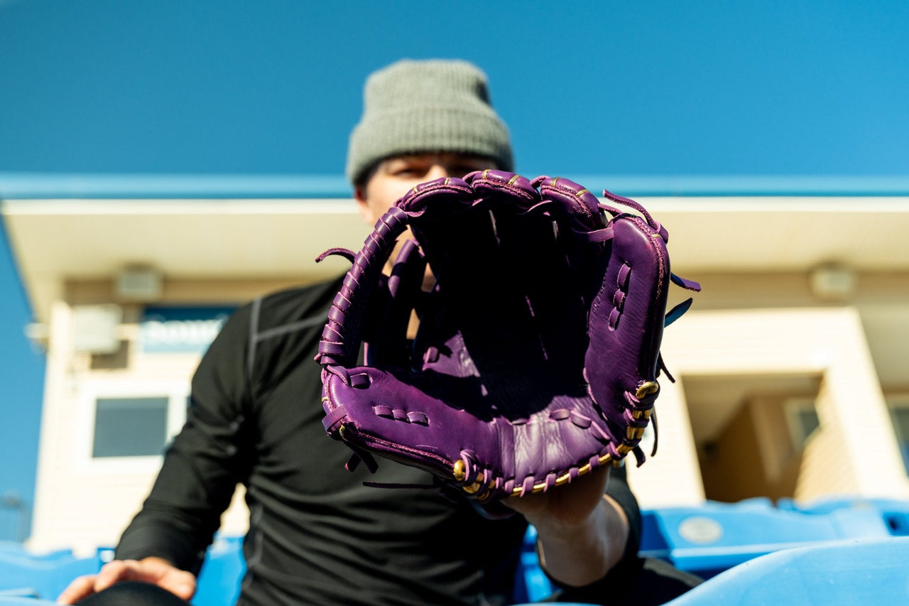 Purple baseball glove with gold lacing held by a person in a gray beanie and black shirt.
