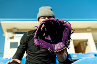 Thumbnail for Purple baseball glove with gold lacing held by a person in a gray beanie and black shirt.