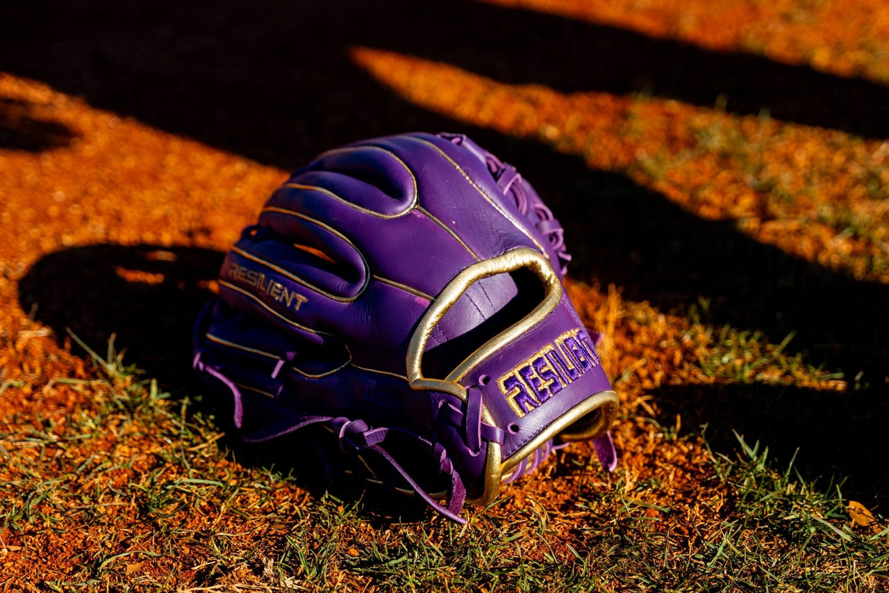 Purple and gold baseball glove on a dirt and grass field, with "RESILIENT" embossed on the palm.