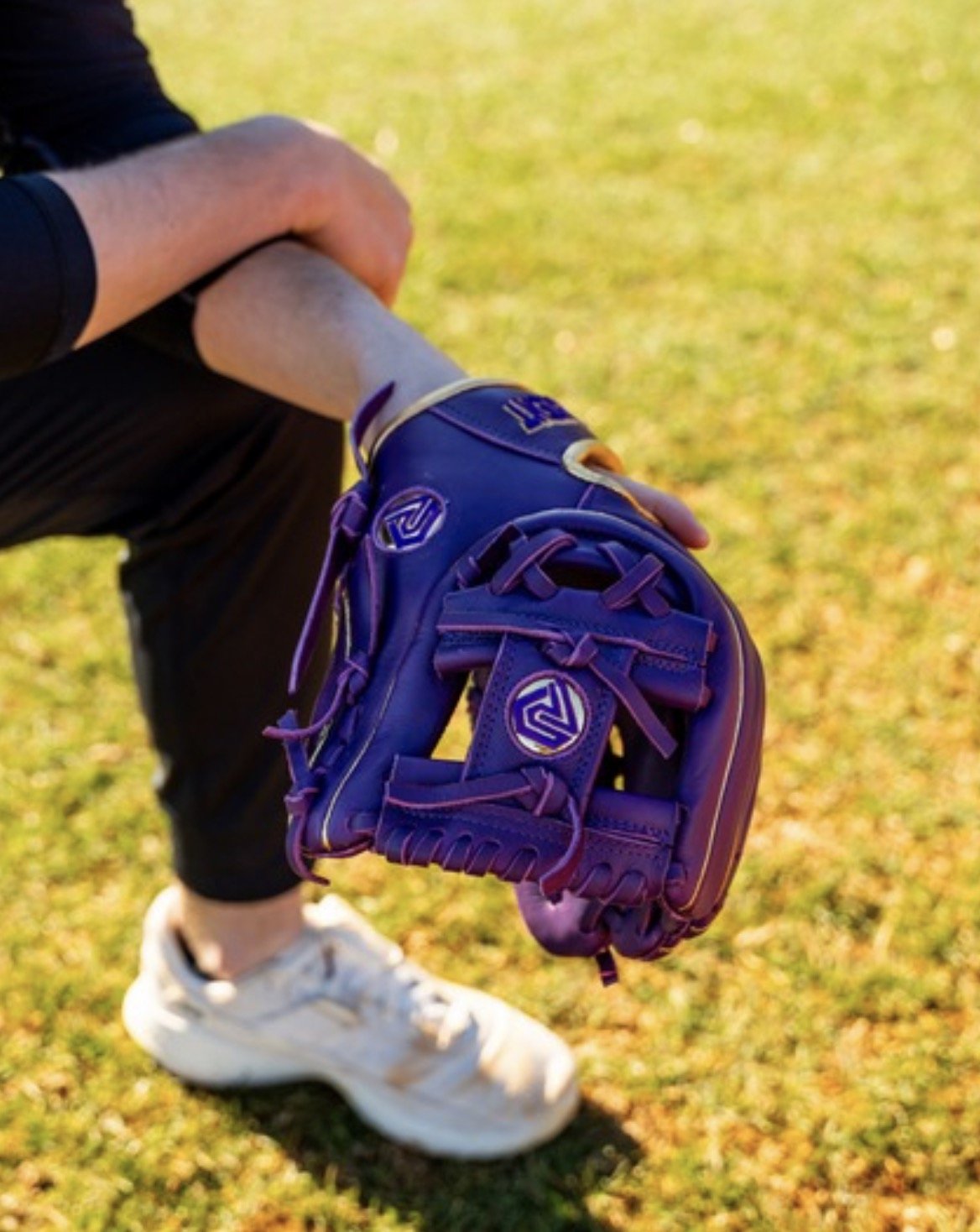 Purple and gold baseball glove on a person's hand, kneeling on green grass.