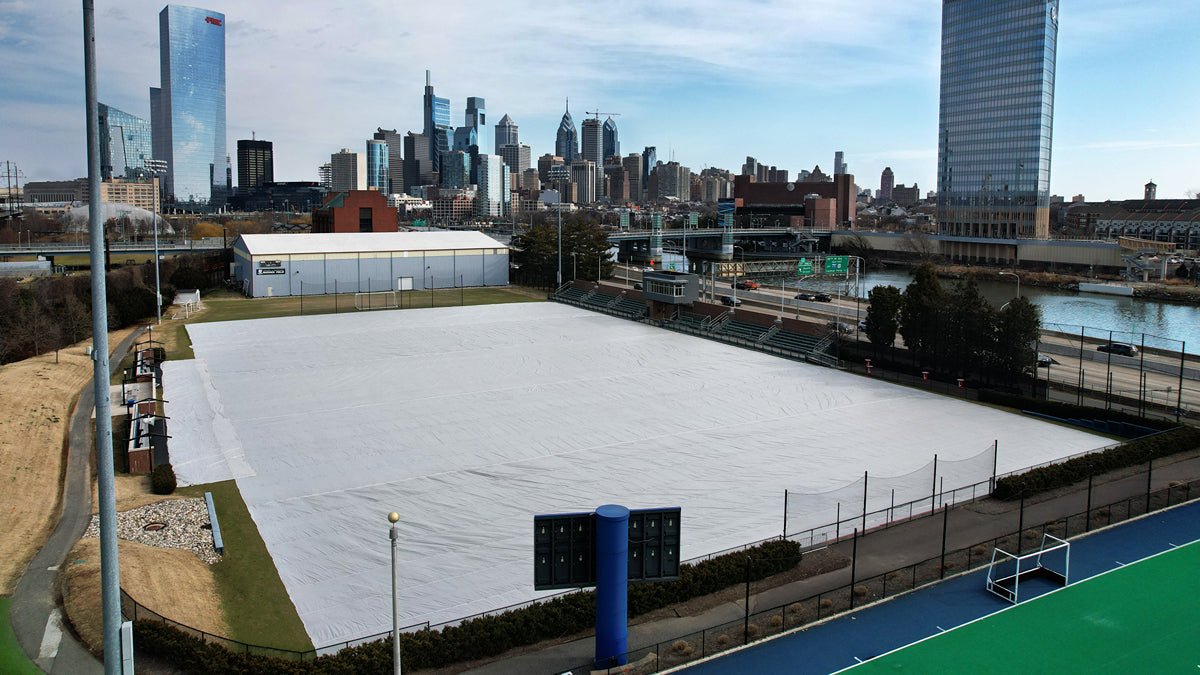 Aerial view of Rhodes Field at the University of Pennsylvania showing a CoverSports growth cover fully deployed across the soccer field in winter, with the Philadelphia skyline including the Comcast Center and PMC Tower visible in the background