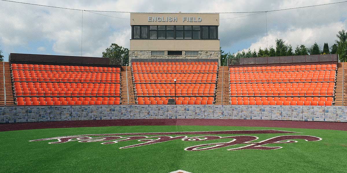 Stone graphic backstop padding below orange stadium seats at a college field