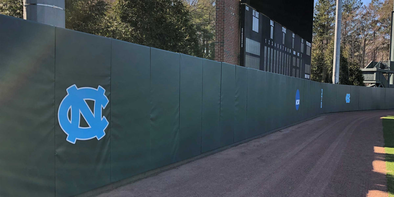 Dark green wood backed outfield wall pads with a school logo running along a baseball warning track