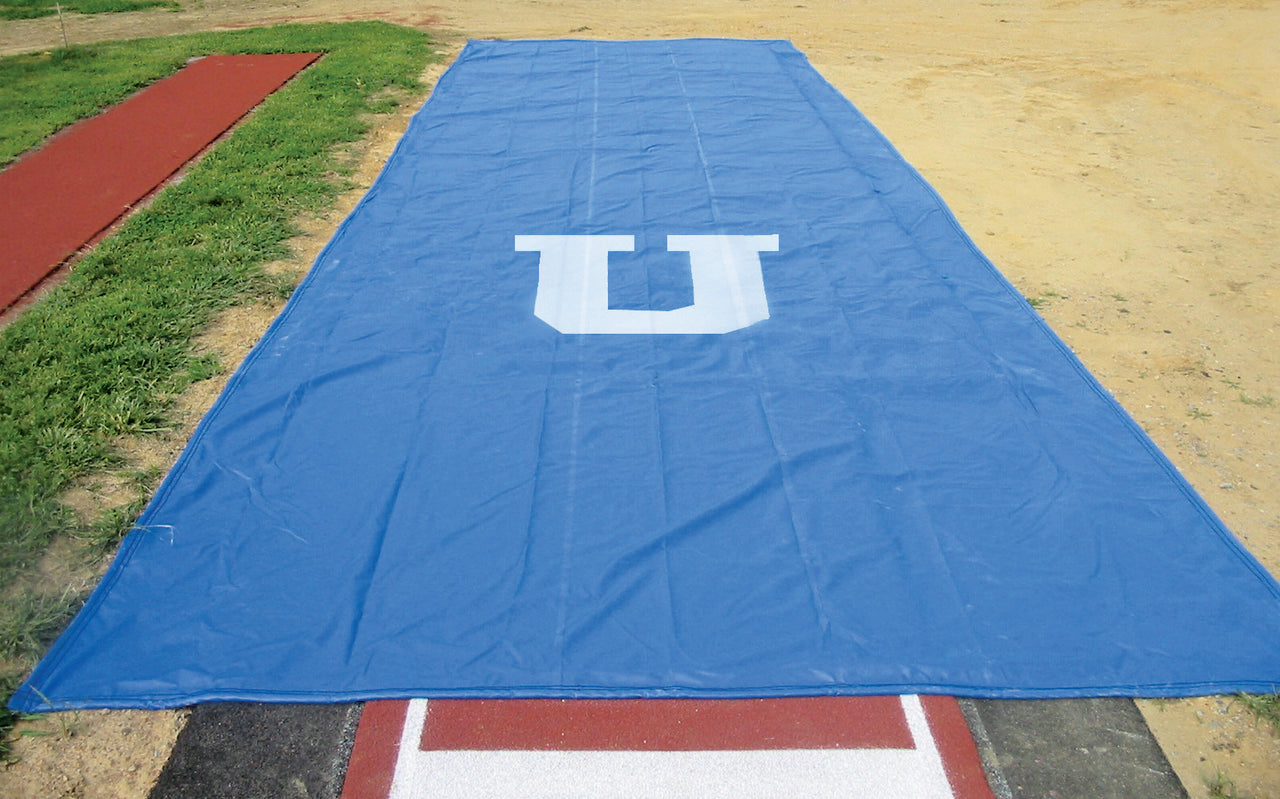 Blue weighted long jump pit cover protecting sand pit with custom printed university logo, showing full installation on outdoor track and field facility